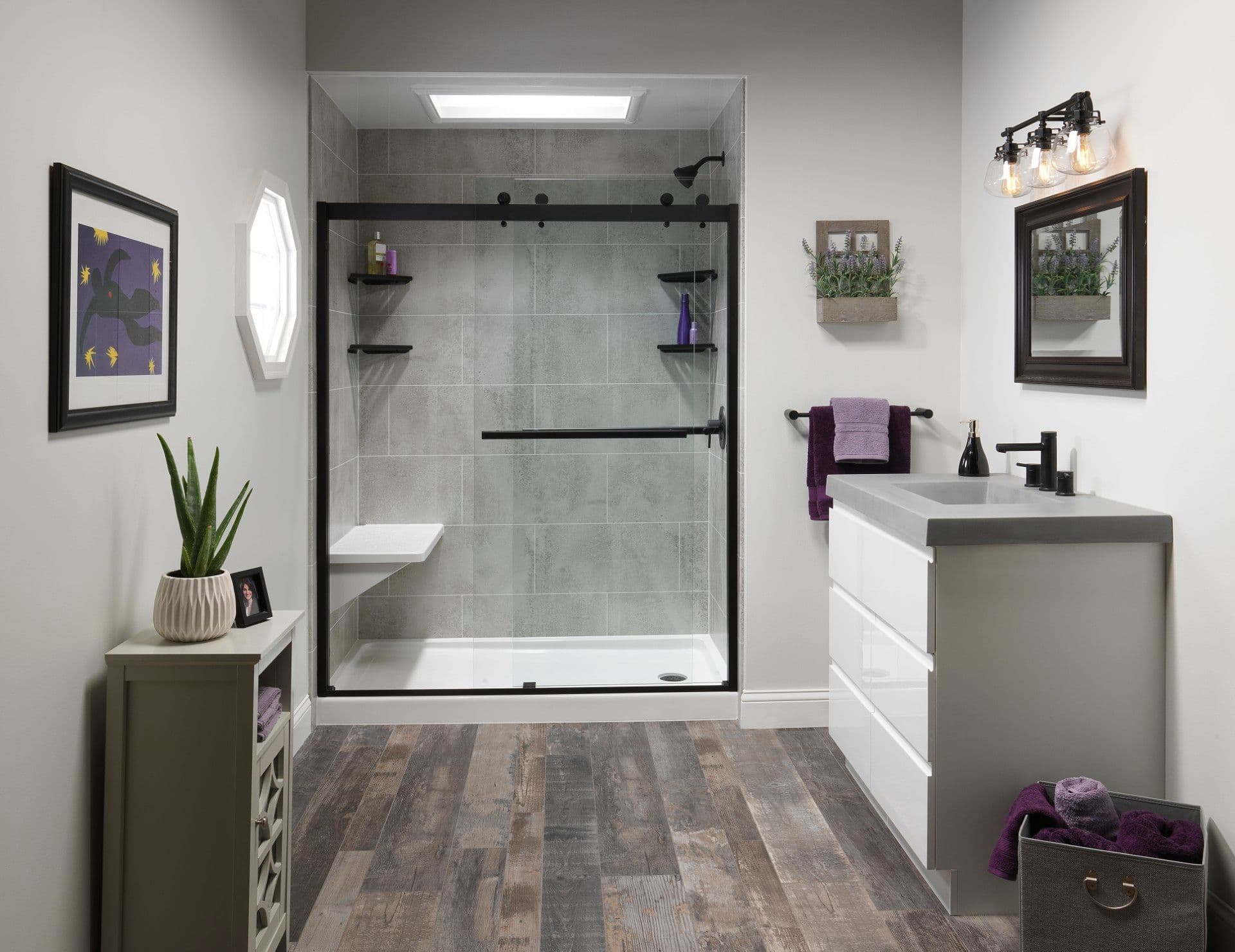 Modern bathroom featuring a spacious walk-in shower with clear sliding glass doors, black hardware, built-in bench and corner shelves, gray tile walls, wood-look flooring, and a sleek white floating vanity with matte black fixtures.