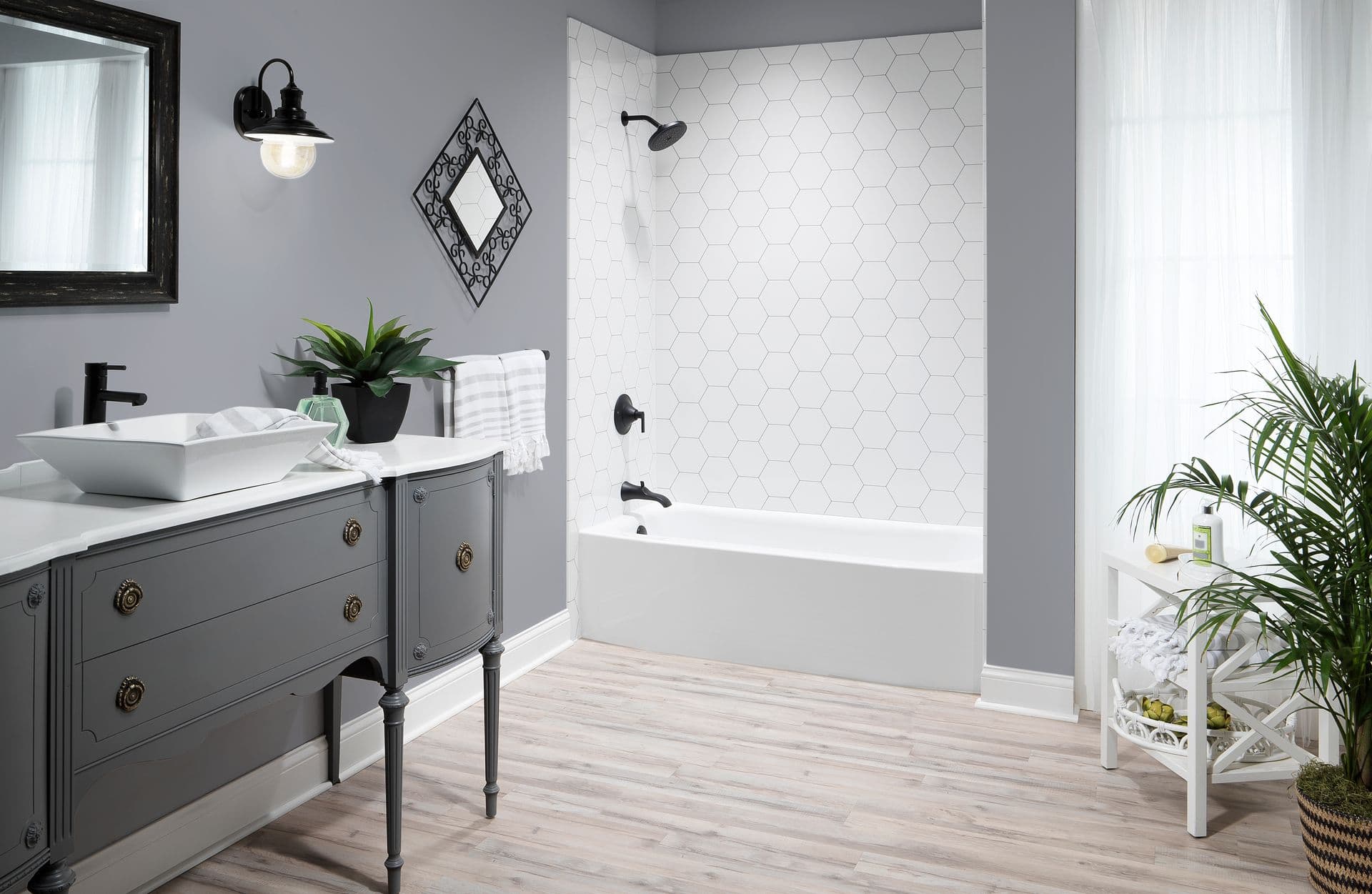 Modern bathroom featuring a white bathtub with a white hexagon tile surround, matte black shower fixtures, a vintage gray vanity with a vessel sink, light wood-look flooring, and bright contemporary décor.