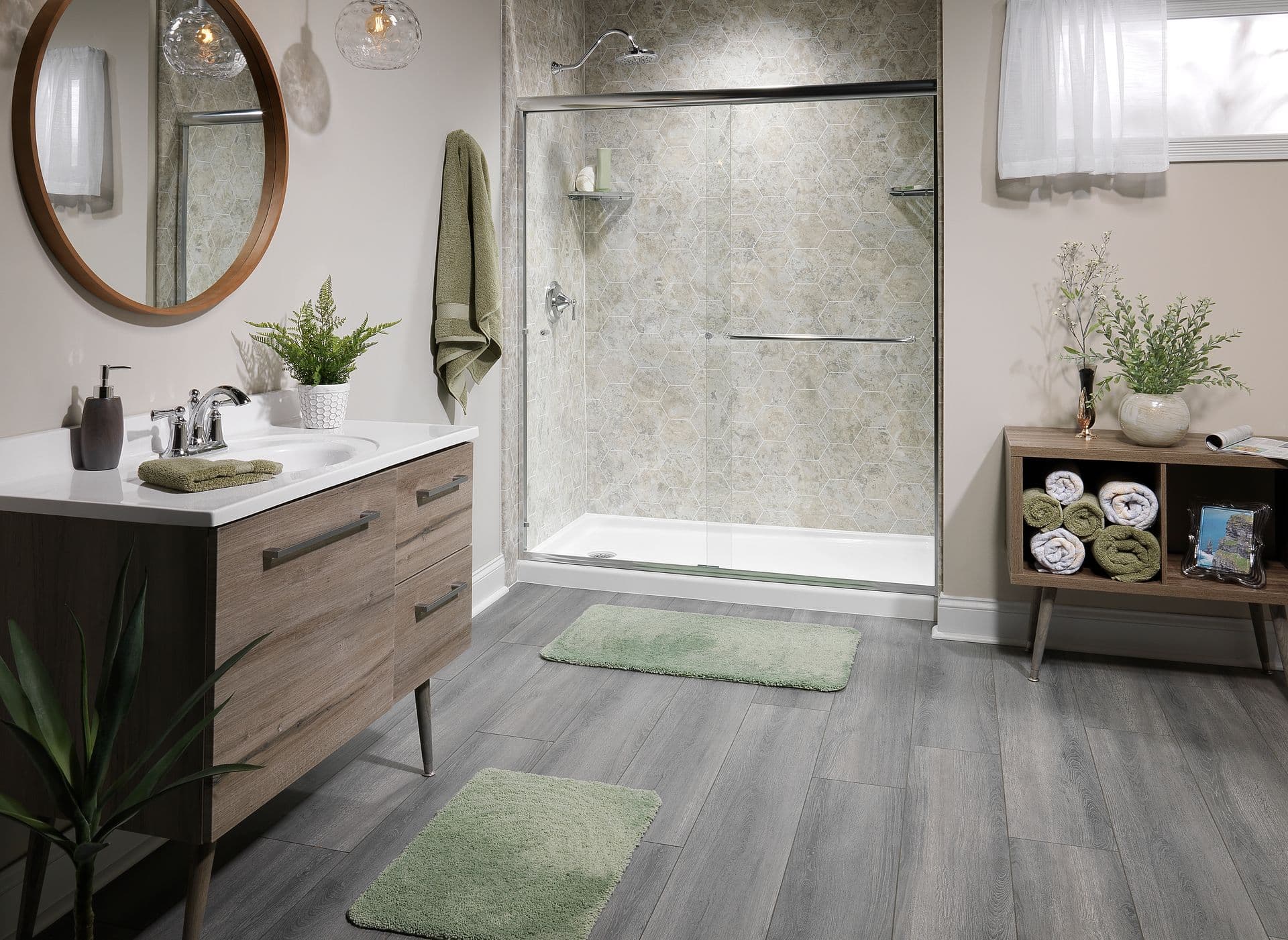 Modern bathroom featuring a frameless sliding glass shower with a gray hexagon-pattern wall surround, wood-look vanity with a white countertop, round wood-framed mirror, gray wood-look flooring, and spa-inspired décor.