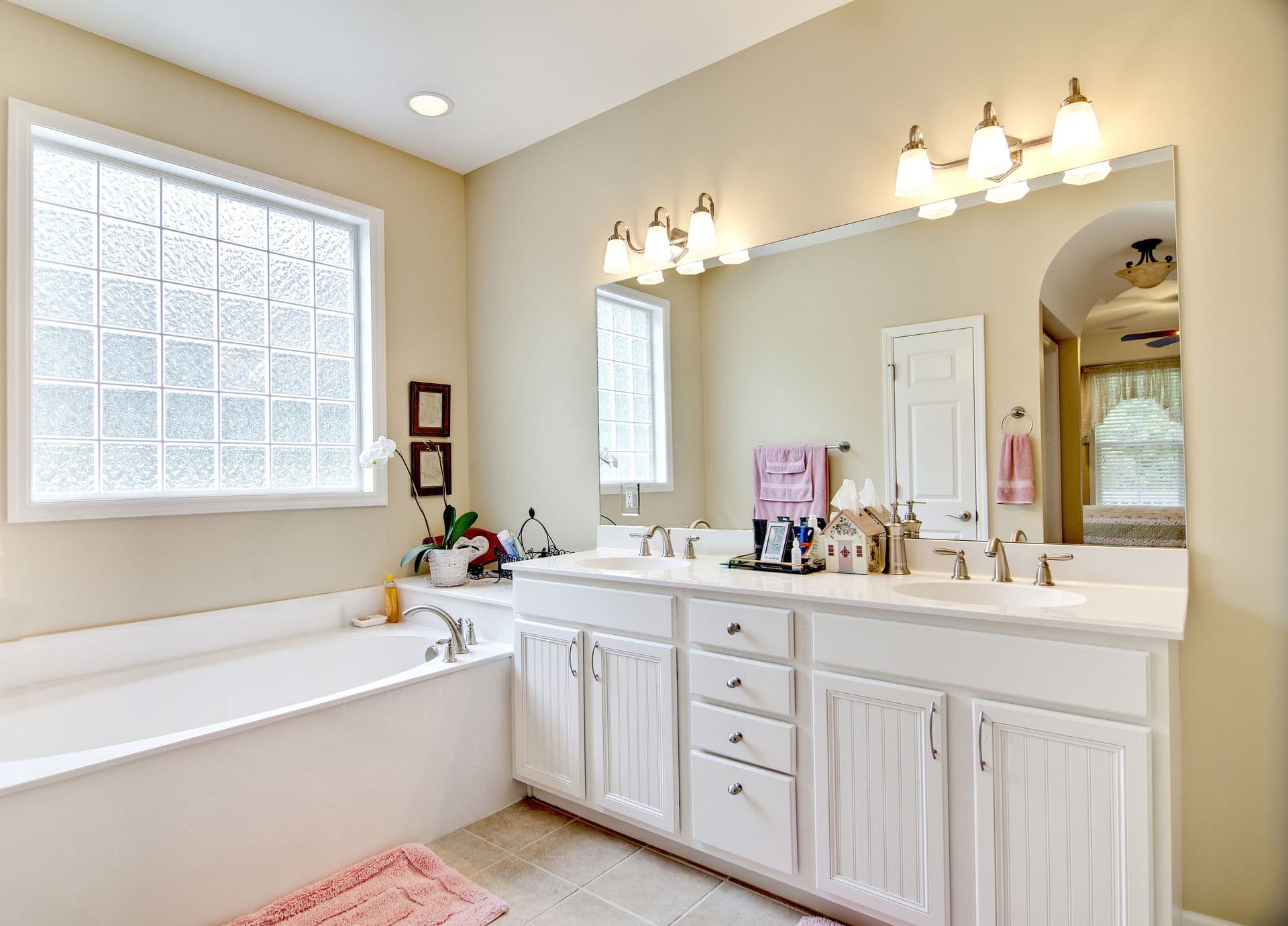 Bright bathroom with a white double-sink vanity, oversized wall mirror, soaking tub beneath a large glass block window, brushed nickel fixtures, beige walls, and light tile flooring.