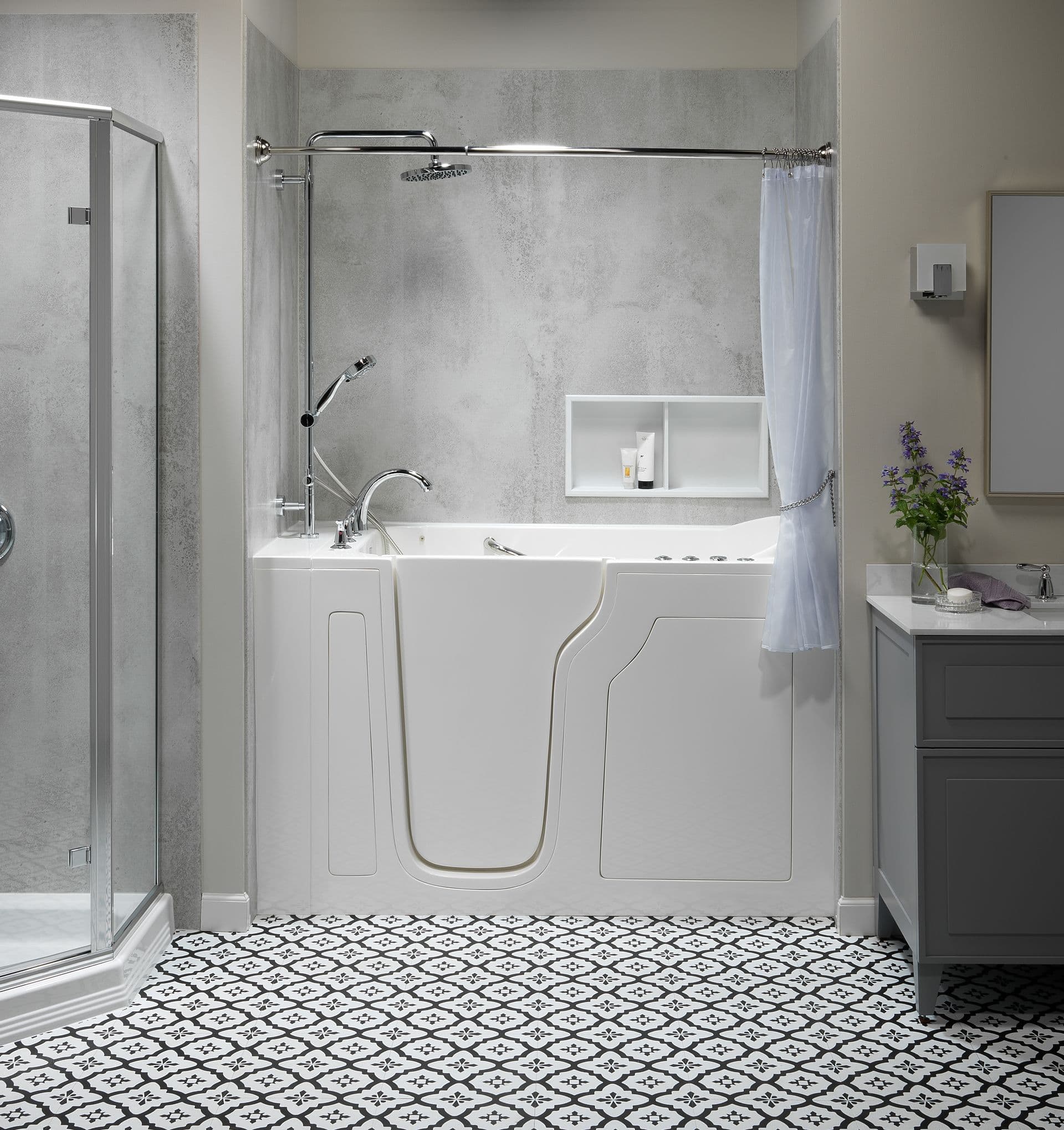 Close-up of a white walk-in tub with a low-entry door, handheld shower, grab bar, built-in shelving niche, and textured gray wall surround.